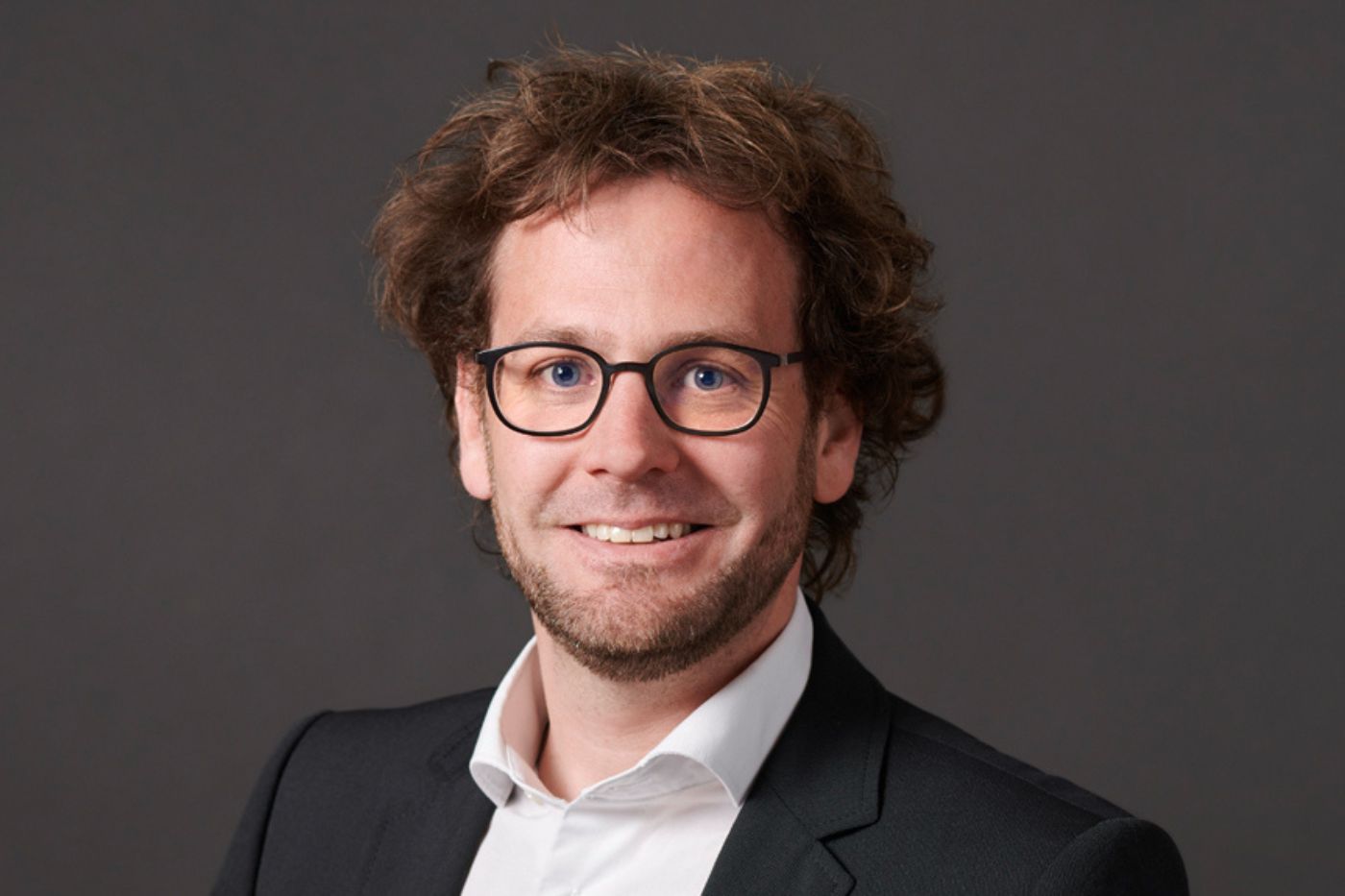 Mann mit lockigem Haar und Brille im dunklen Anzug vor neutralem Hintergrund lächelnd porträtiert.