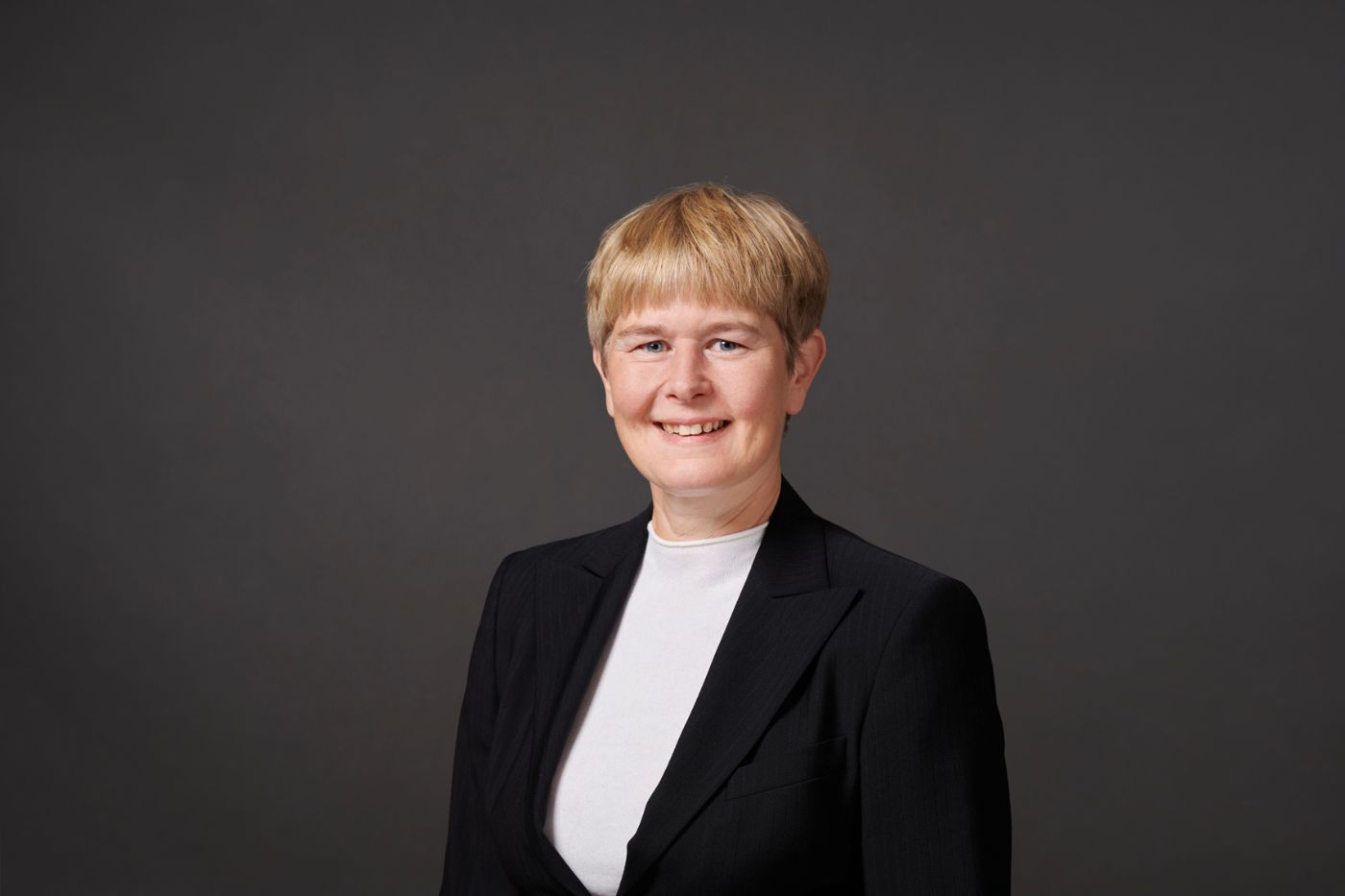 Frau mit kurzem blondem Haar in dunklem Blazer und hellem Oberteil vor neutralem Hintergrund lächelnd porträtiert.
