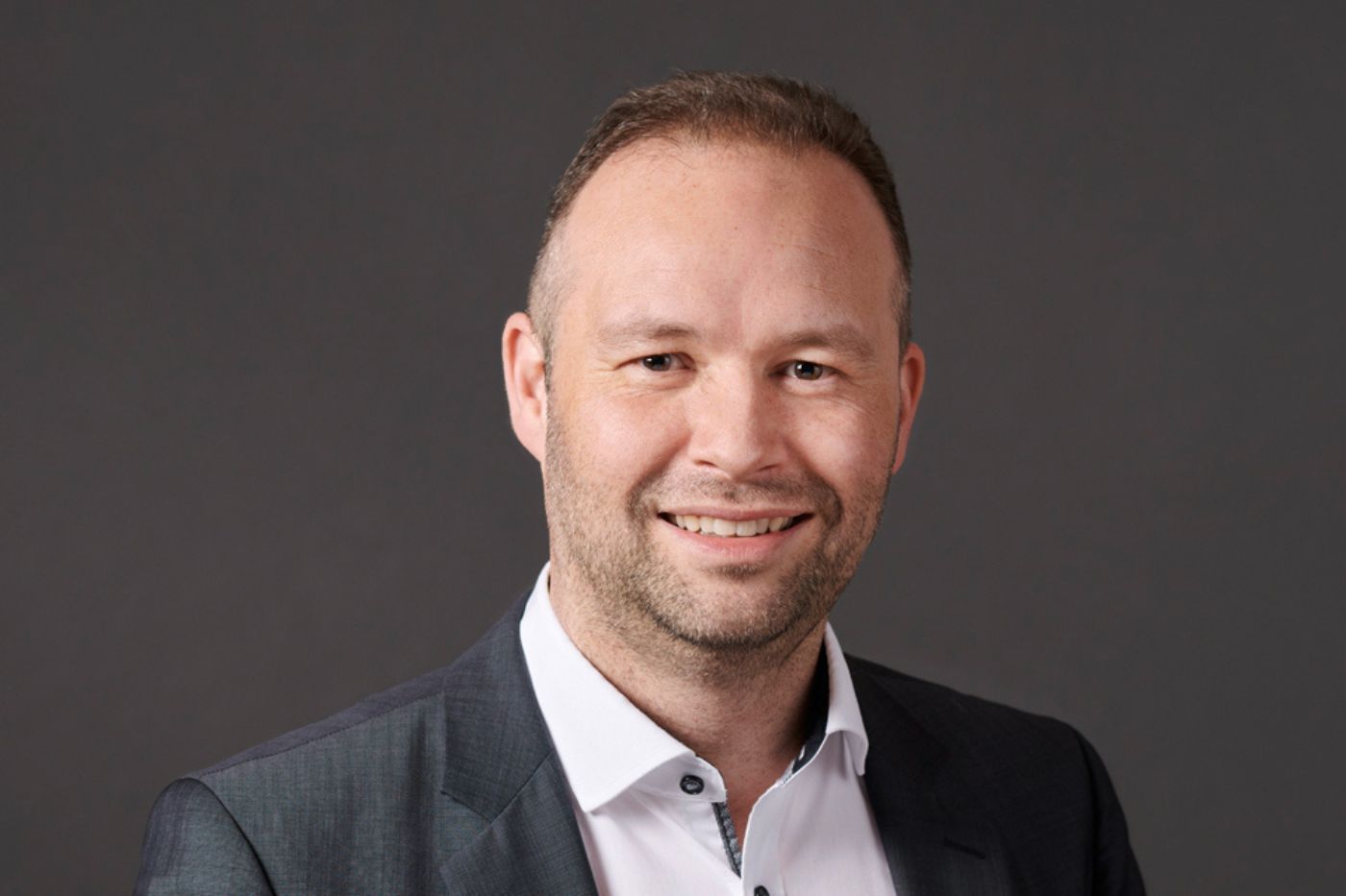 Mann mit kurzem Haar im dunklen Anzug und weißem Hemd vor neutralem Hintergrund lächelnd porträtiert.