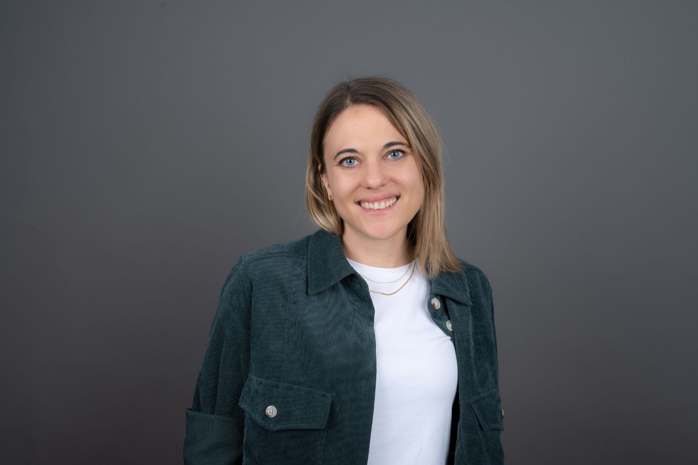 Frau mit schulterlangem blondem Haar in blauer Cordjacke und weißem Shirt vor neutralem Hintergrund lächelnd porträtiert.