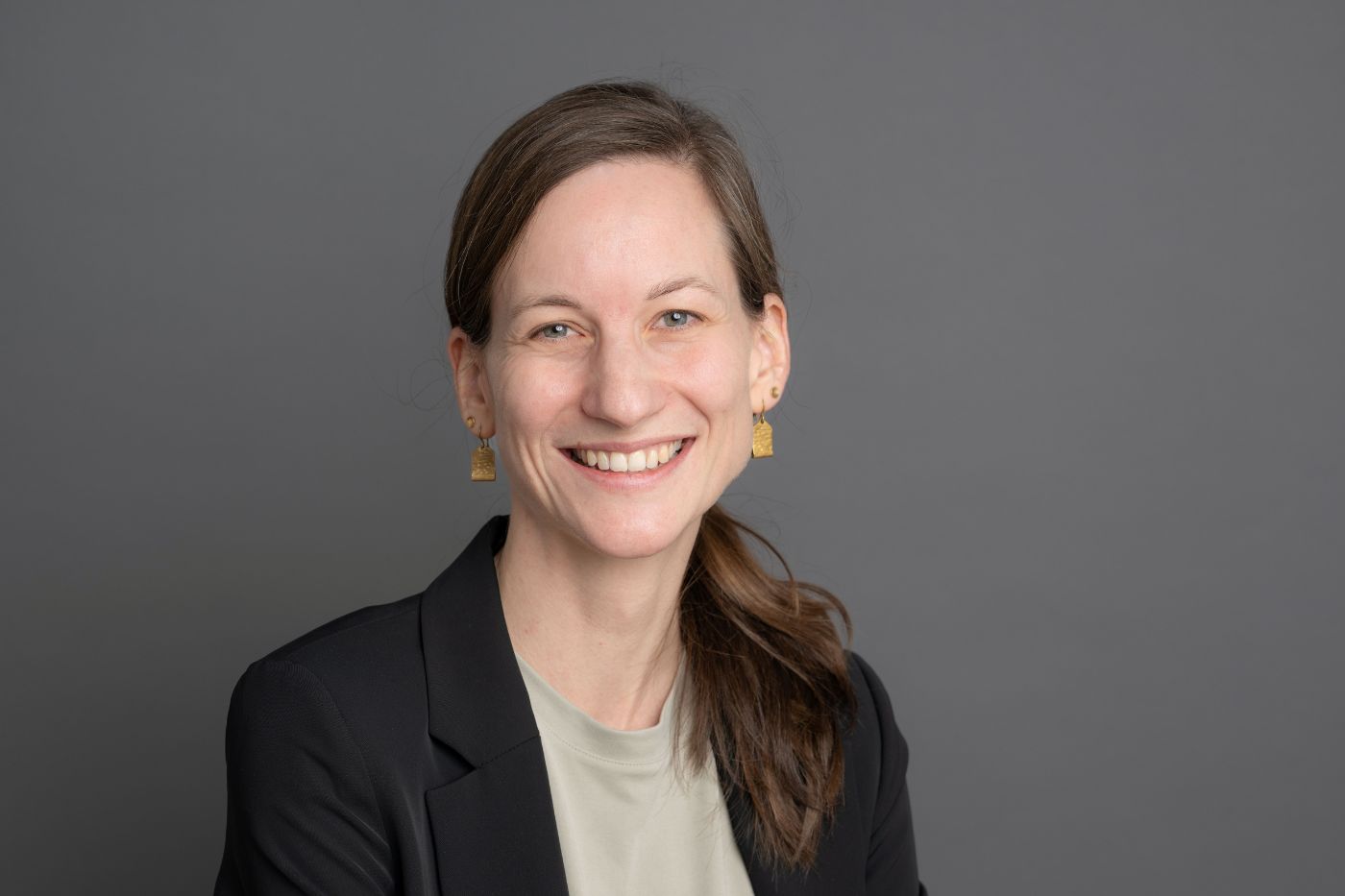 Frau mit seitlich gebundenem braunem Haar in dunklem Blazer und hellem Shirt vor neutralem Hintergrund lächelnd porträtiert.