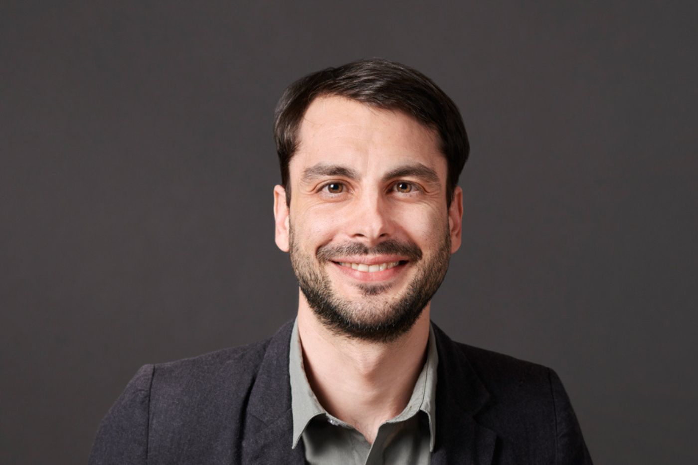 Mann mit kurzem dunklem Haar und Bart im dunklen Sakko vor neutralem Hintergrund lächelnd porträtiert.