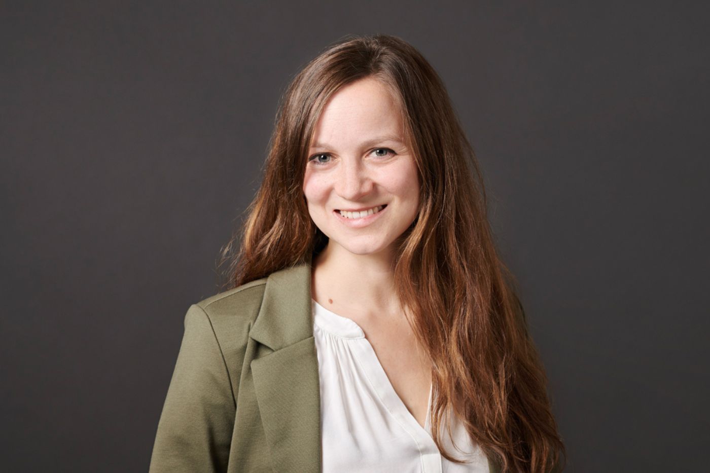 Frau mit langen braunen Haaren in olivgrünem Blazer und weißer Bluse vor neutralem Hintergrund lächelnd porträtiert.