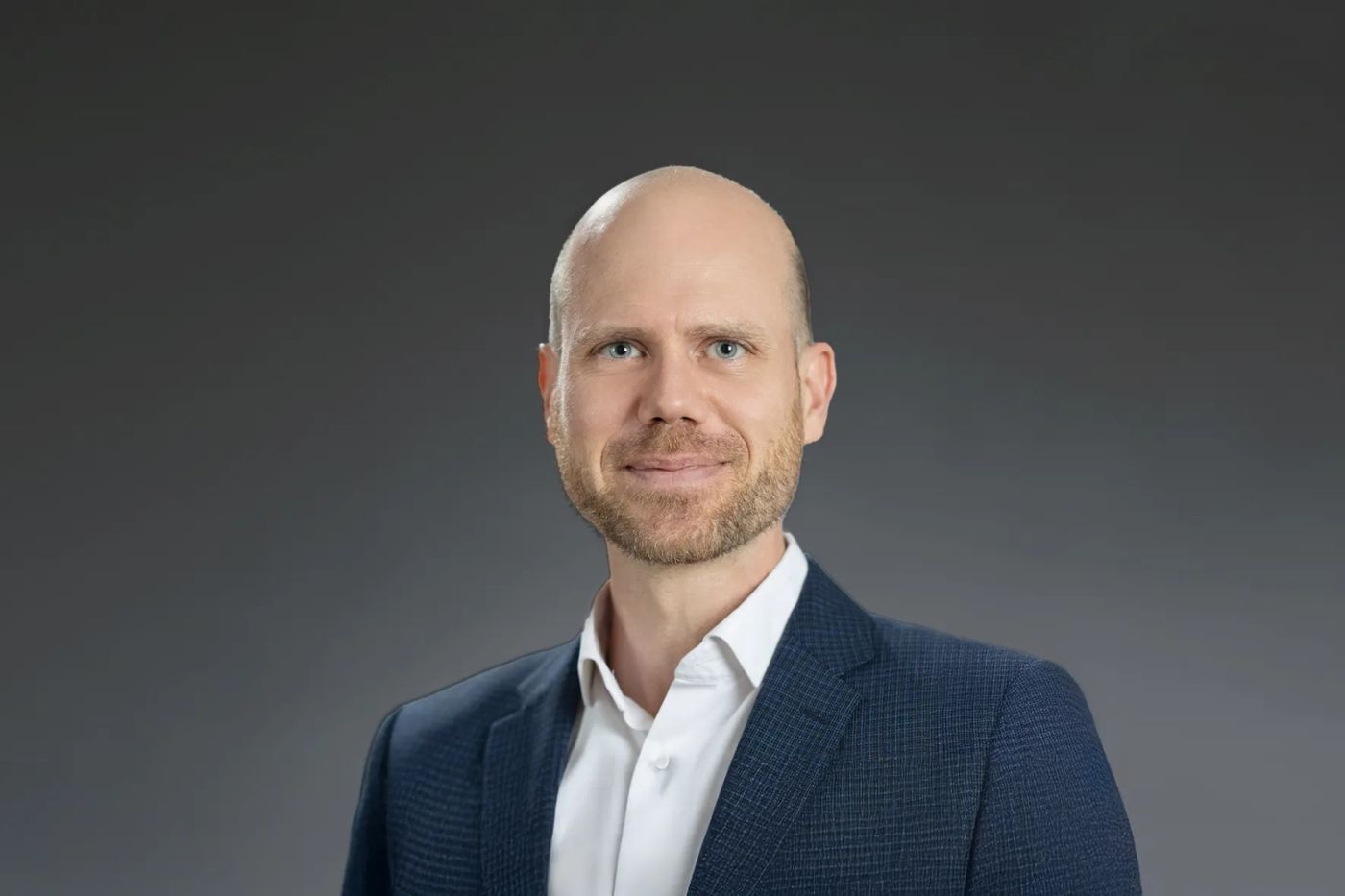 Mann mit Glatze und kurzem Bart im dunkelblauen Sakko und weißem Hemd vor neutralem Hintergrund lächelnd porträtiert.