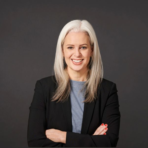 Frau mit langen blonden Haaren lächelt im Porträt vor dunklem Hintergrund und trägt einen Blazer.