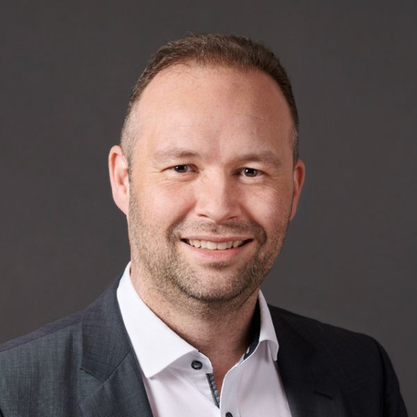 Mann mit kurzem Haar und Sakko lächelt im Porträt vor dunklem Hintergrund.