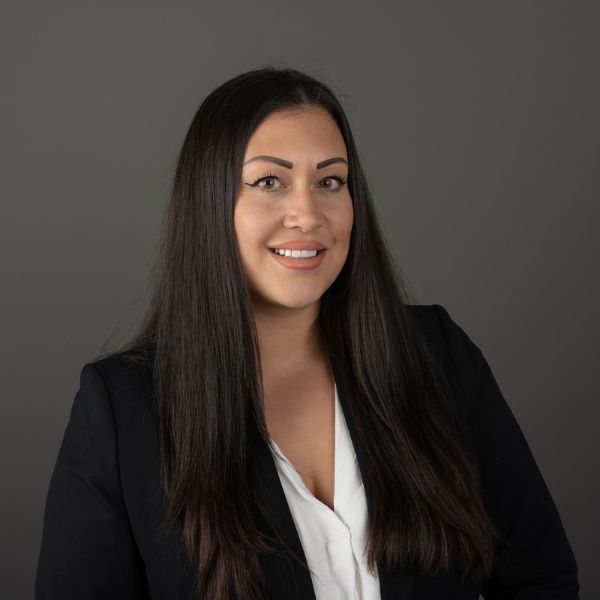 Frau mit langen dunklen Haaren lächelt im Porträt vor dunklem Hintergrund und trägt Blazer und Bluse.