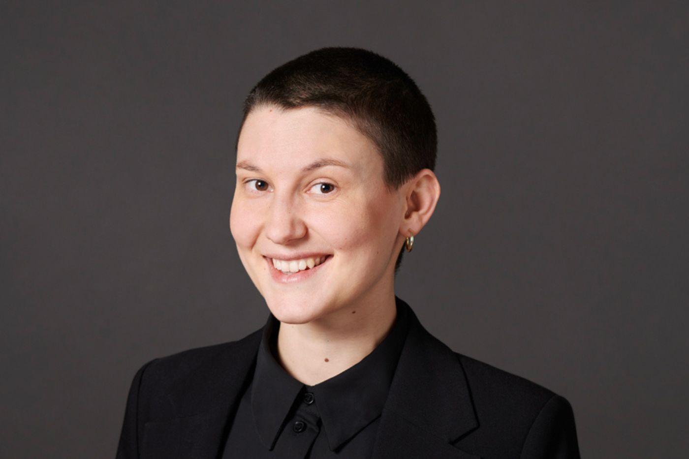 Frau mit sehr kurzem Haar in dunklem Blazer und schwarzem Hemd vor neutralem Hintergrund lächelnd porträtiert.